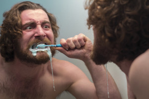 Silly bearded man brushing teeth in bathroom mirror